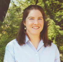 Ruth Anderson, woman with long brown hair, smiling in front of evergreen trees wearing a light blue shirt