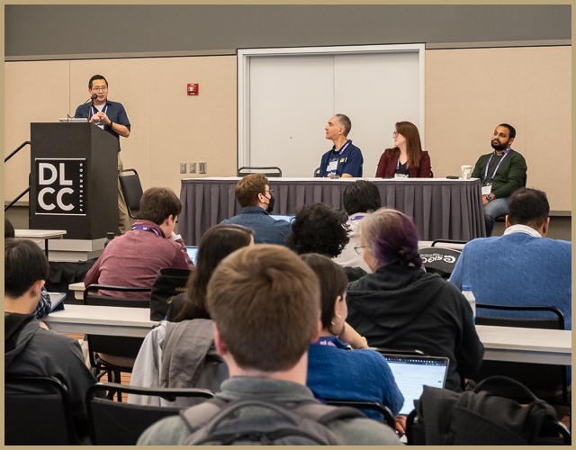 Justin standing at a podium, speaking as part of a panel of four professors to an audience