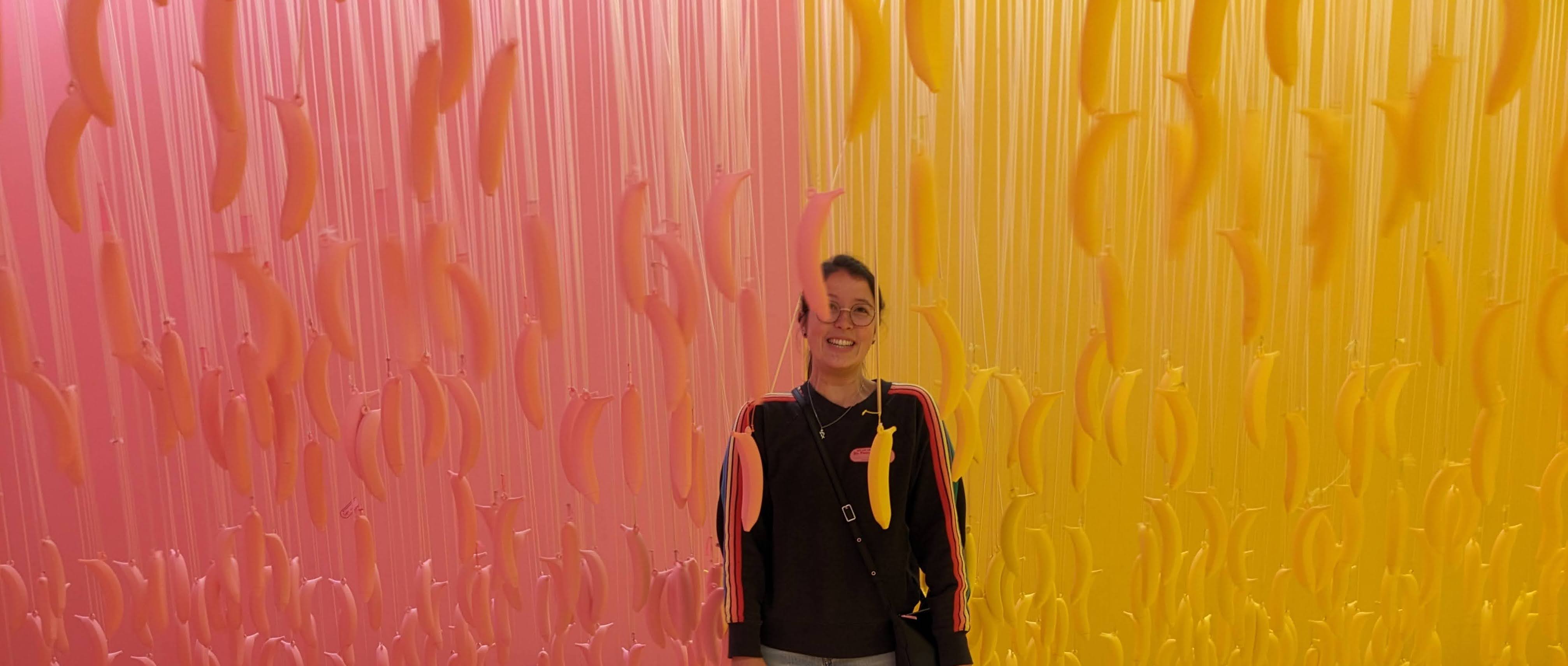 half-asian female a ponytail and glasses standing in front of a half-pink, half-yellow room with bananas hanging from strings in matching colors