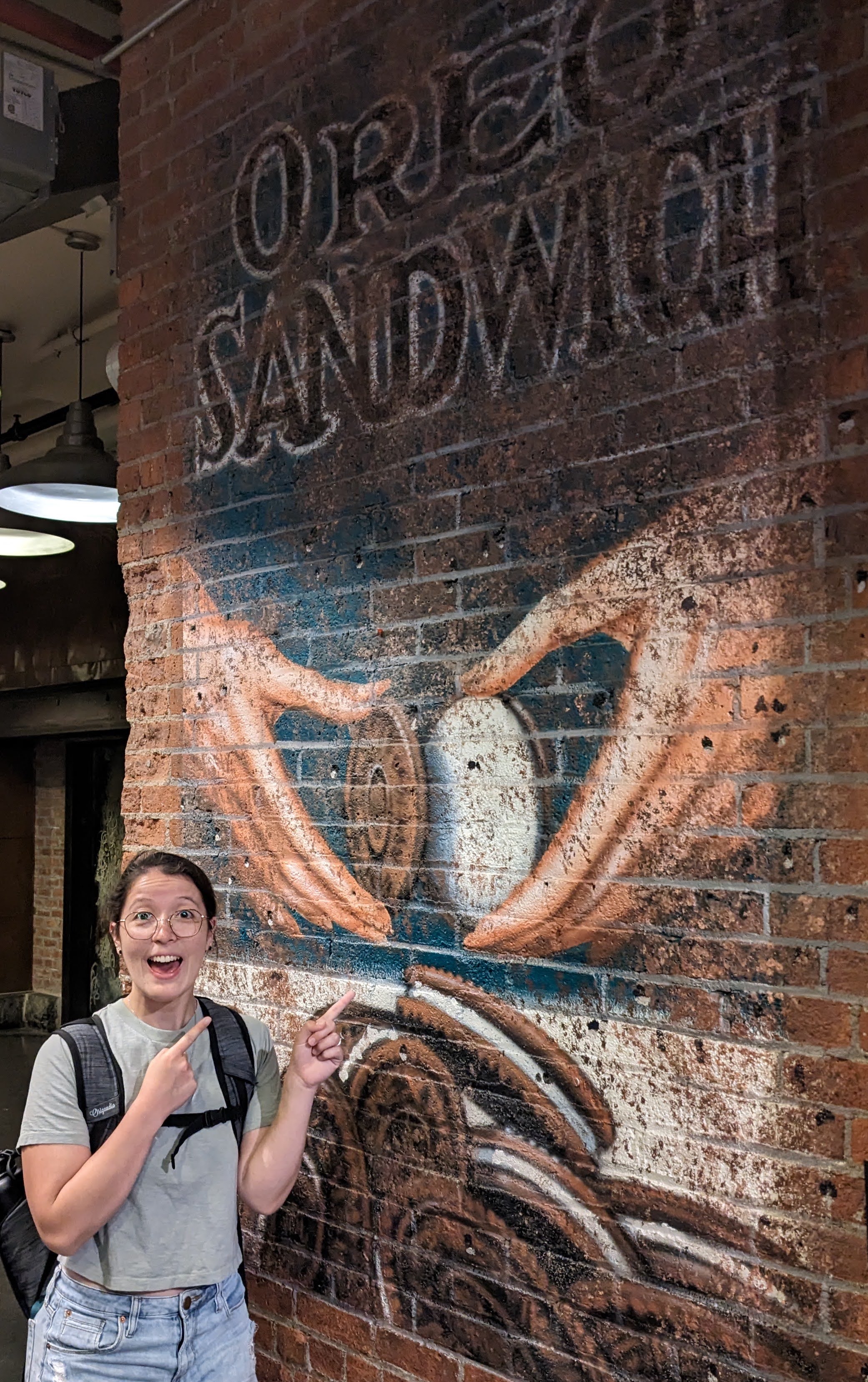 half-asian female with a ponytail and glasses pointing to a mural of an old oreo sandwich cookie advertisement on a brick wall