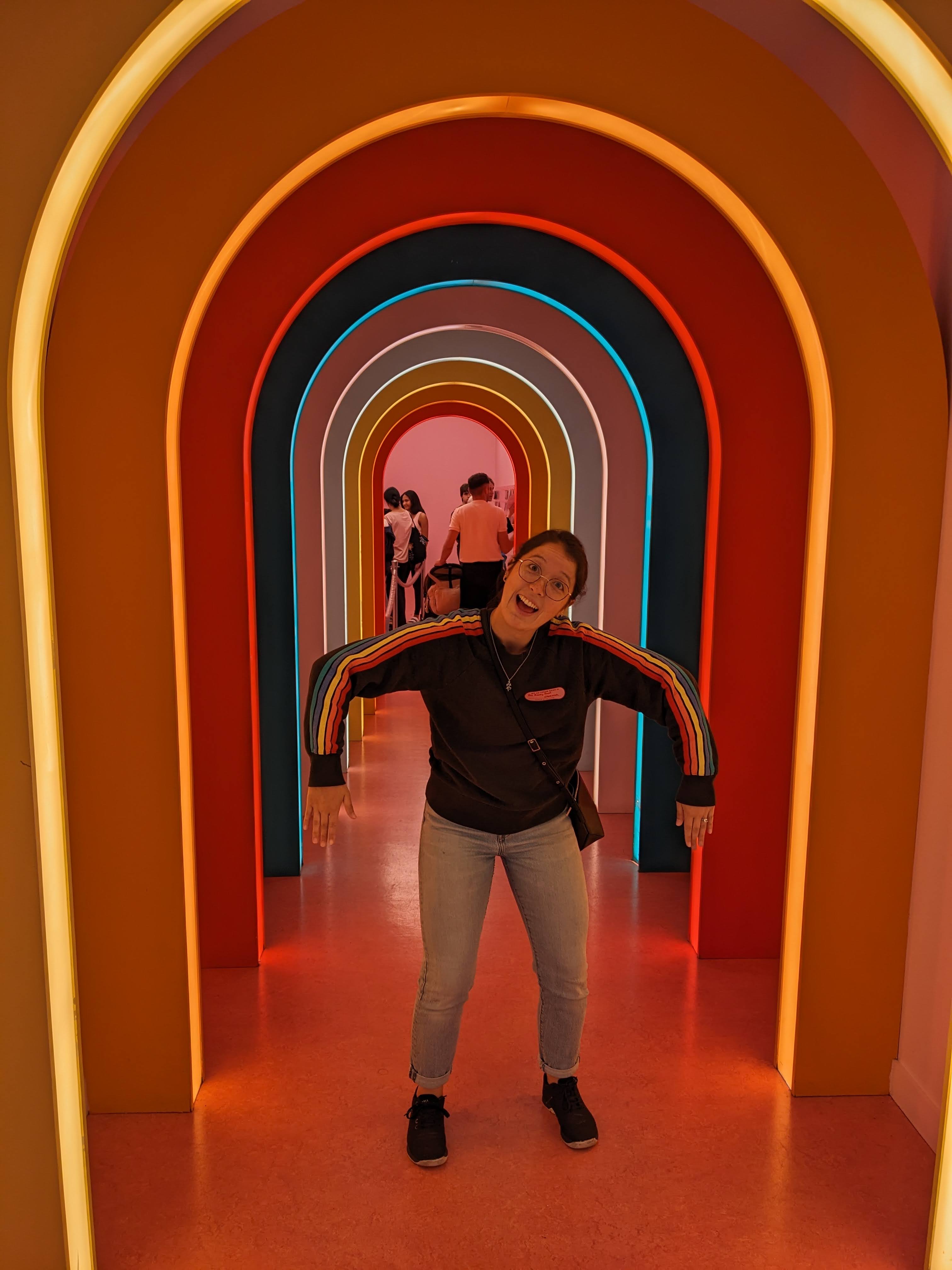 half-asian female standing in a tunnel of neon lights wearing a sweatshirt with rainbow lines down the sleeves with arms making a rainbow shape